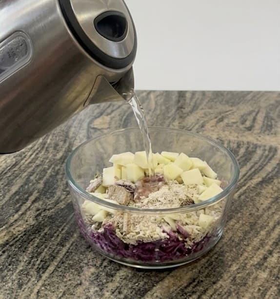 Hot water pouring from a kettle into a bowl of oats, cabbage, and diced apples.